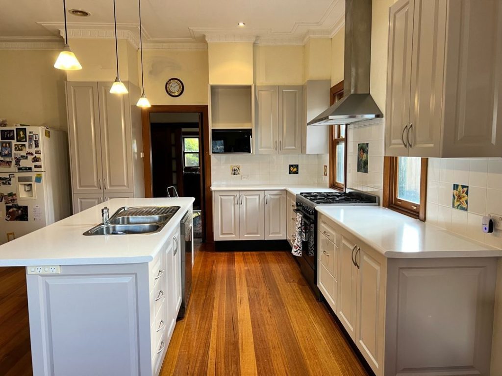 A modern kitchen with a central island, stainless steel appliances, white cabinetry, wooden flooring, and pendant lighting.