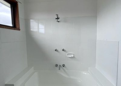 White-tiled bathroom with a bathtub, overhead showerhead, faucet handles, and natural light from a window.