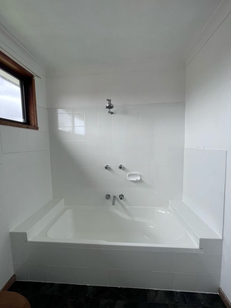 White-tiled bathroom with a bathtub, overhead showerhead, faucet handles, and natural light from a window.