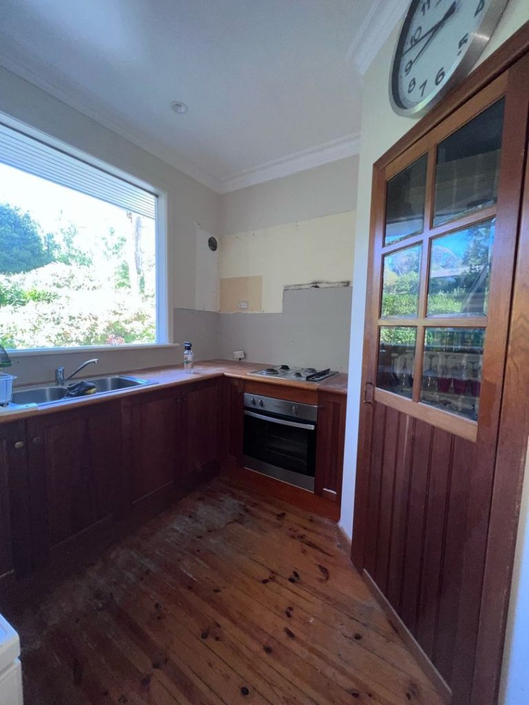 Kitchen interior with wooden cabinets, stainless steel sink, electric stove, large window, and wall clock above a wooden door.