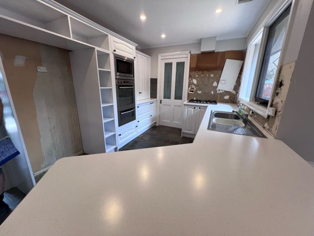 Kitchen under renovation with white cabinets, built-in oven and microwave, stovetop, double sink, and exposed wall patches.