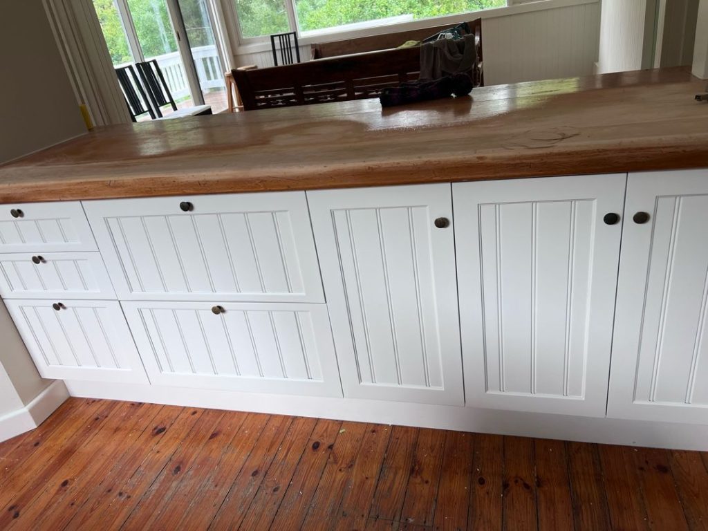 White kitchen cabinets with a wooden countertop in a room with hardwood floors and large windows.