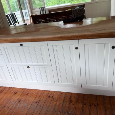 White kitchen cabinets with a wooden countertop in a room with hardwood floors and large windows.