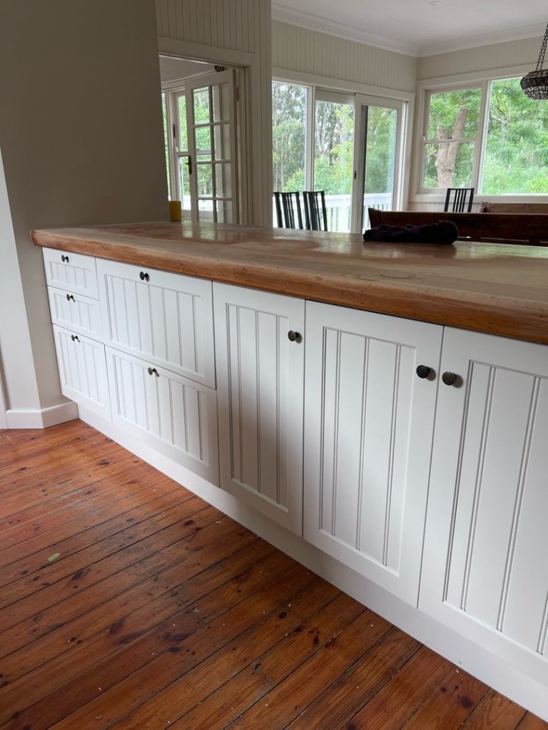 White kitchen cabinets with a wooden countertop in a room with large windows and a view of greenery.