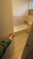 Person opening door to an outdated bathroom with beige tiles and a stained wall above the bathtub.