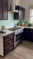 Modern kitchen with dark wooden cabinets, stainless steel oven and range hood, light green backsplash, and various countertop items.