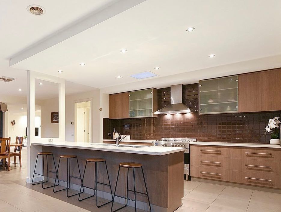 Modern kitchen with island, bar stools, wood-finished cabinets, stainless steel range hood, and brown tile backsplash.