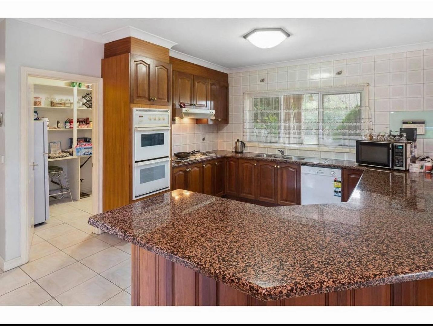 A kitchen with an L-shaped granite countertop, wooden cabinets, double oven, stovetop, range hood, microwave, and visible pantry.