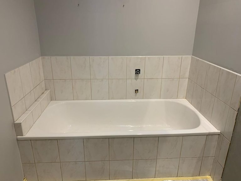 A white bathtub surrounded by new white tiles with a subtle marble pattern in a light gray painted bathroom.