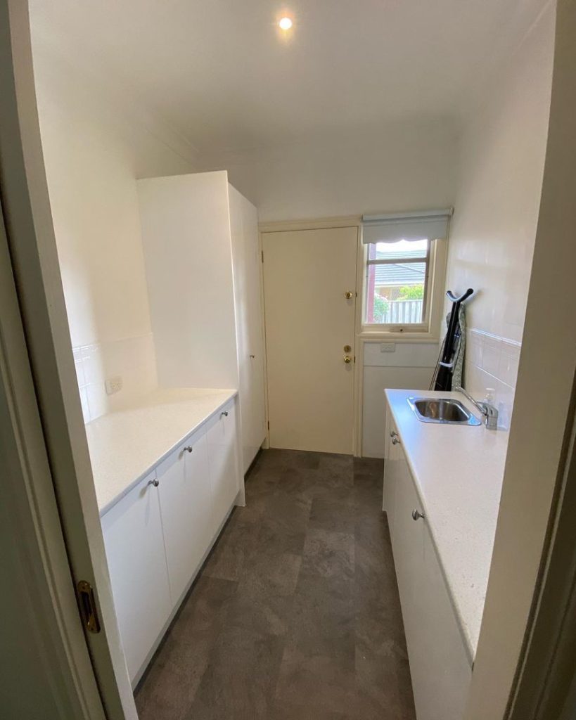 A modern laundry room with white cabinets, countertops, a stainless steel sink, an ironing board, and gray tiled flooring.