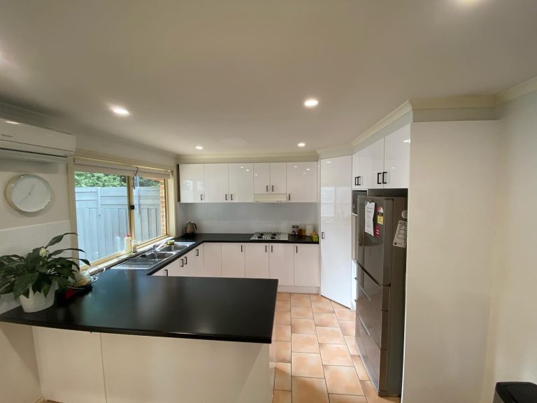 Modern kitchen with white cabinets, black countertops, stainless steel refrigerator, window view of greenery, and recessed lighting.