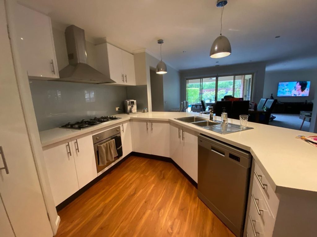 Modern kitchen with white cabinets, stainless steel appliances, wooden flooring, and an open-plan layout extending to a living room.