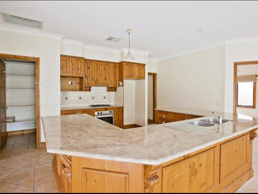 An outdated kitchen with wooden cabinets, a central island with a sink, a gas stove, and an open pantry.