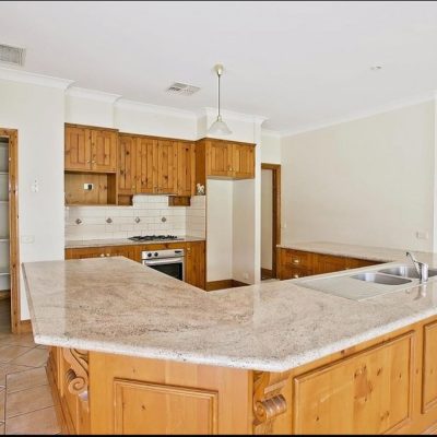 An outdated kitchen with wooden cabinets, a central island with a sink, a gas stove, and an open pantry.