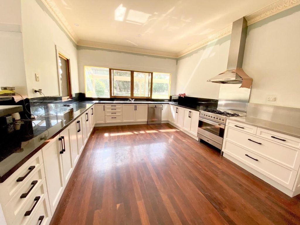 Modern kitchen featuring white cabinetry with black handles, black countertops, a large stainless steel stove and range hood, and wooden flooring.