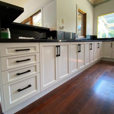 Modern kitchen with white cabinets, black countertops, and wooden floors illuminated by natural light from large windows.