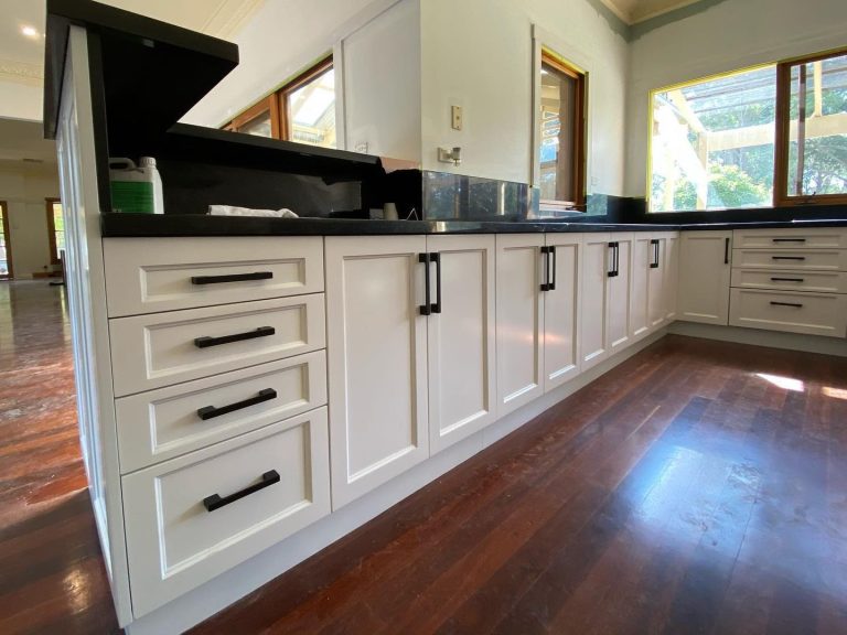 Modern kitchen with white cabinets, black countertops, and wooden floors illuminated by natural light from large windows.