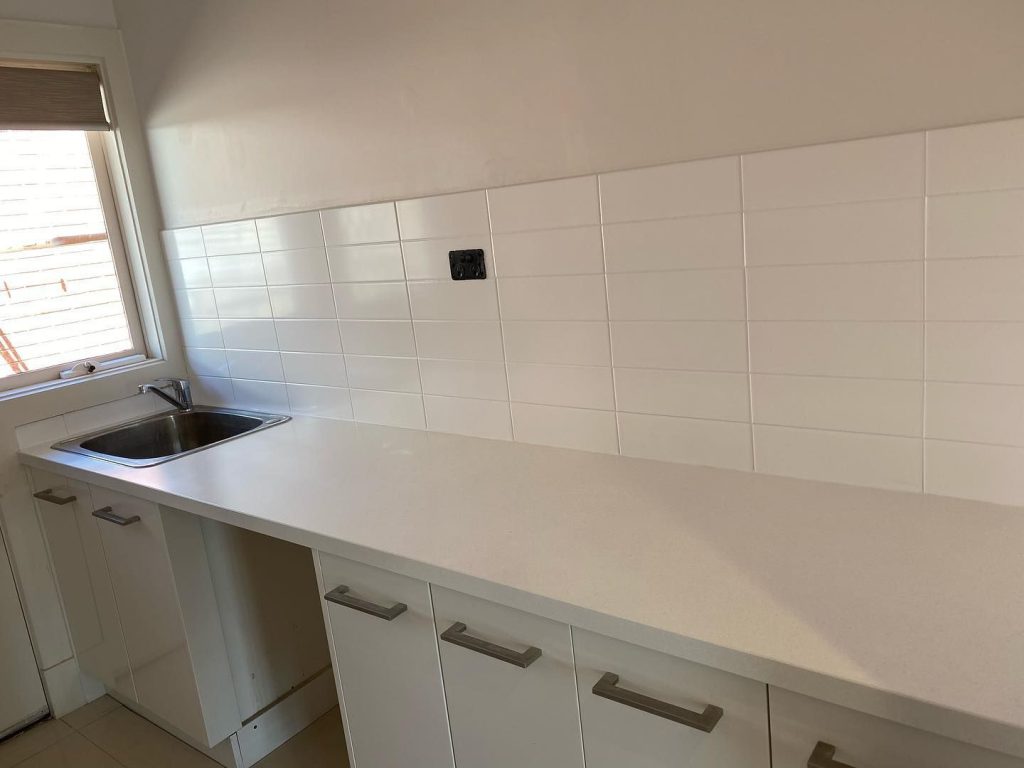 Resurfaced kitchen area with white countertop, stainless steel sink, white tiled backsplash, and white cabinetry.