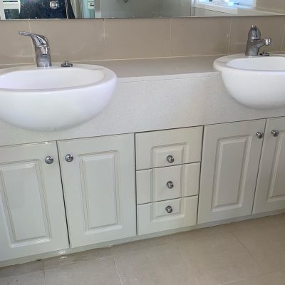 Bathroom vanity with two white sinks, chrome faucets, beige tile backsplash, and light-colored cabinetry.