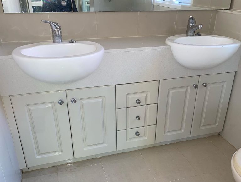 Bathroom vanity with two white sinks, chrome faucets, beige tile backsplash, and light-colored cabinetry.