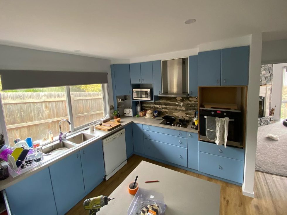 Modern kitchen with blue cabinets, stainless steel appliances, large window, and wooden floor.