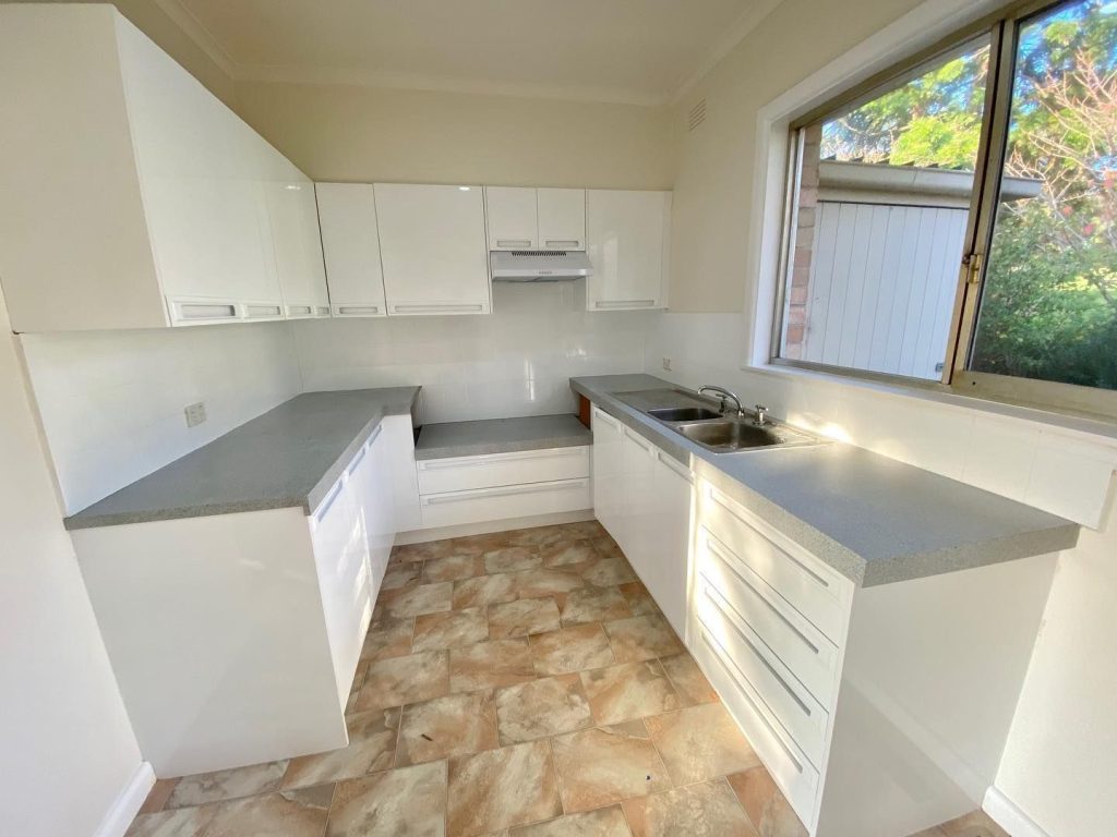 Small renovated kitchen with white cabinets, grey countertops, double sink, and large window.