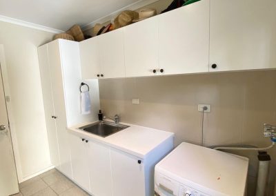 Image of a laundry room with a washing machine, sink, and white storage cabinets topped with various hats.