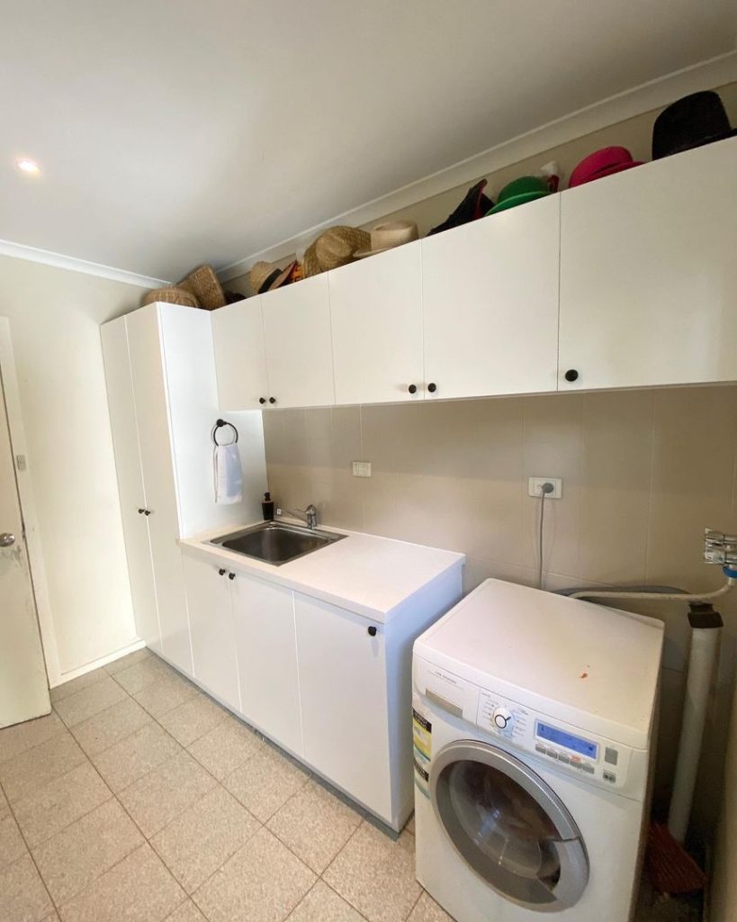 Image of a laundry room with a washing machine, sink, and white storage cabinets topped with various hats.