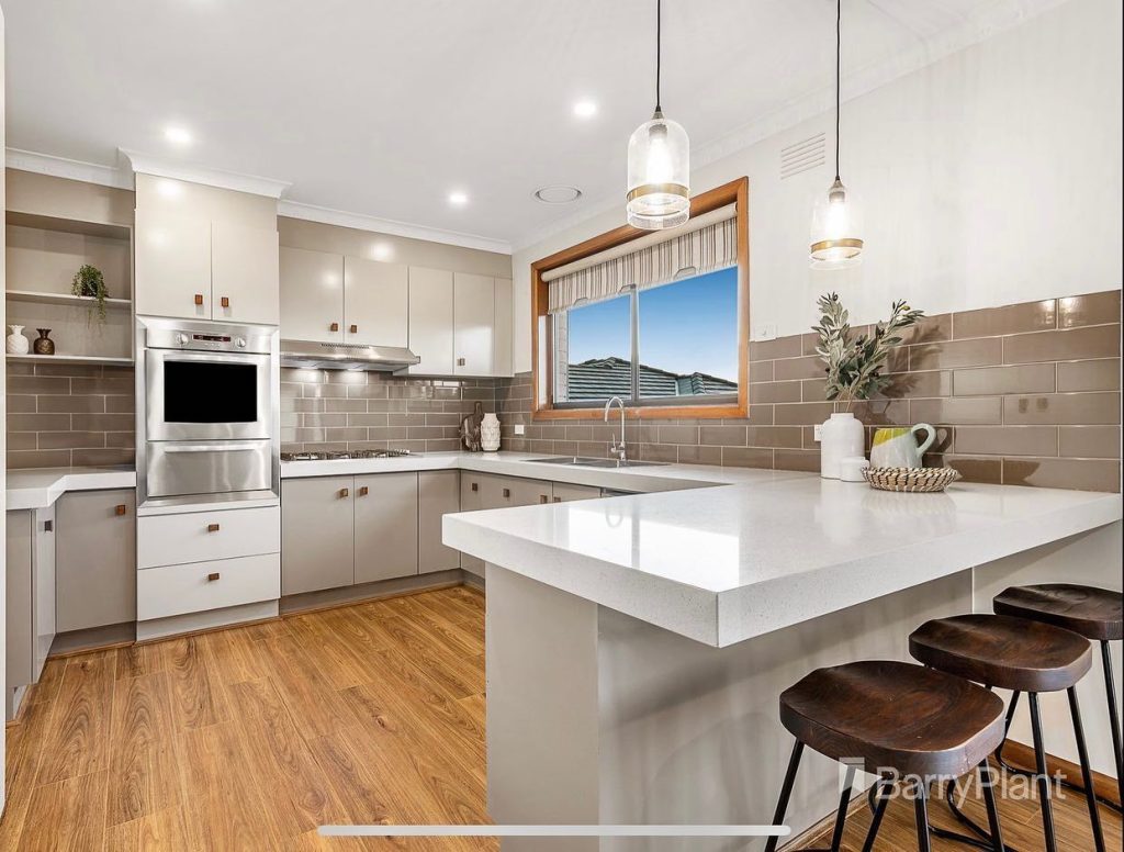 Modern kitchen with white countertops, brown cabinets, stainless steel appliances, wooden flooring, pendant lights, and decorative items.