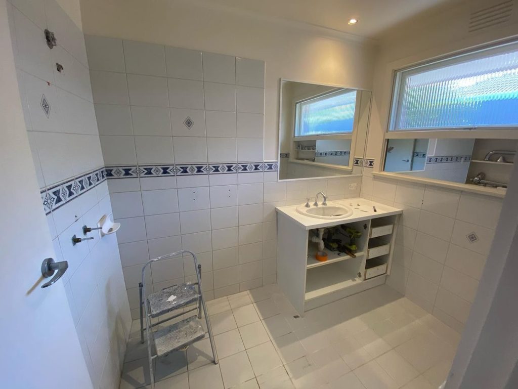 Partially renovated bathroom with white tiles, decorative border, open sink vanity, and step ladder.