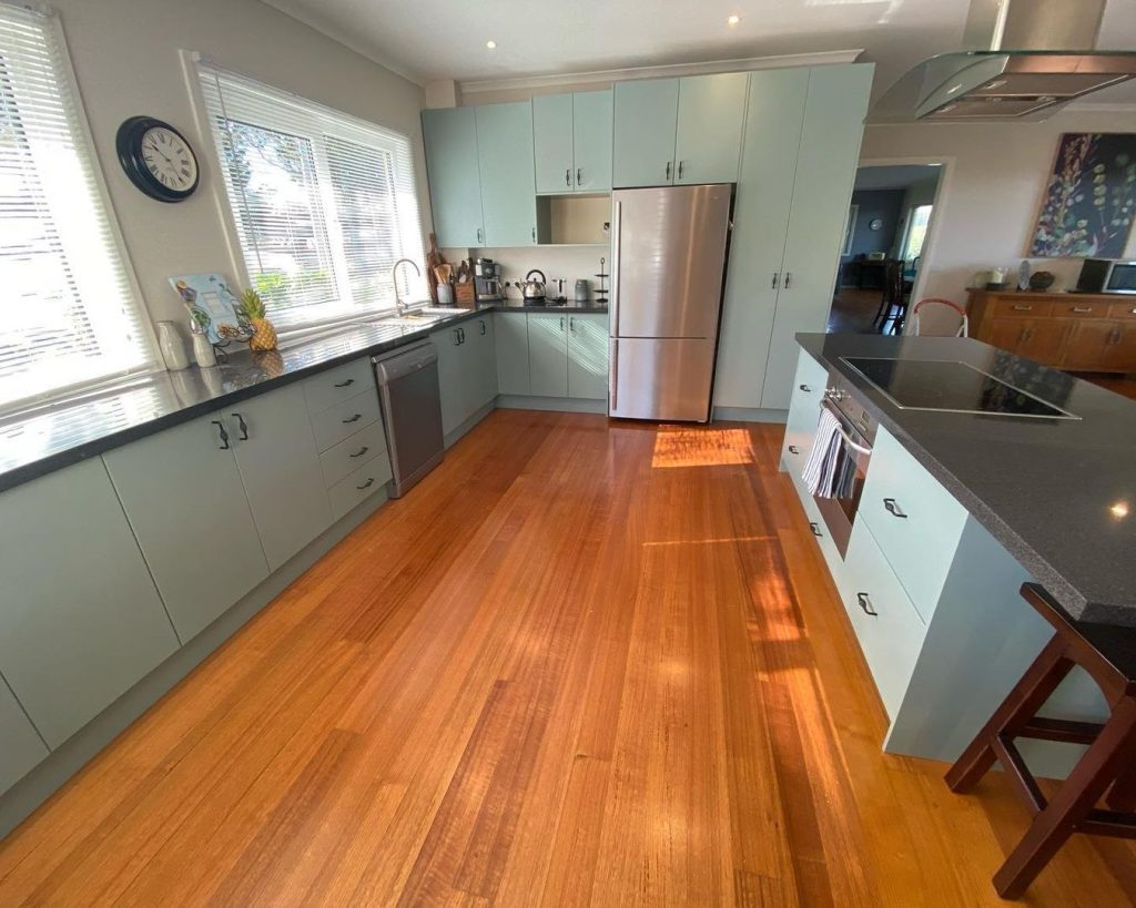 Modern kitchen with light blue cabinets, stainless steel appliances, black countertops, wooden flooring, and large windows.