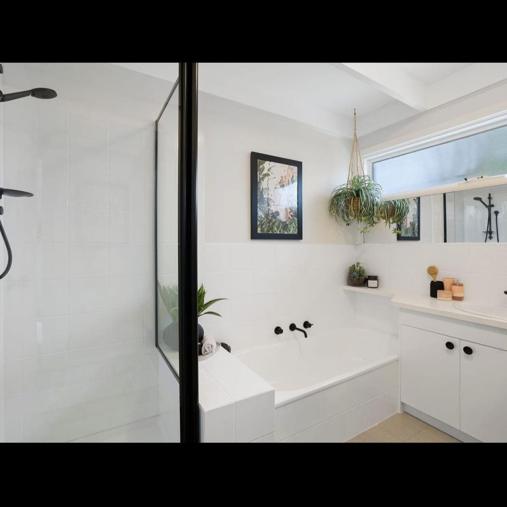 Modern bathroom with white tiles, black fixtures, glass shower enclosure, bathtub, framed picture, hanging plant, and decorative items.