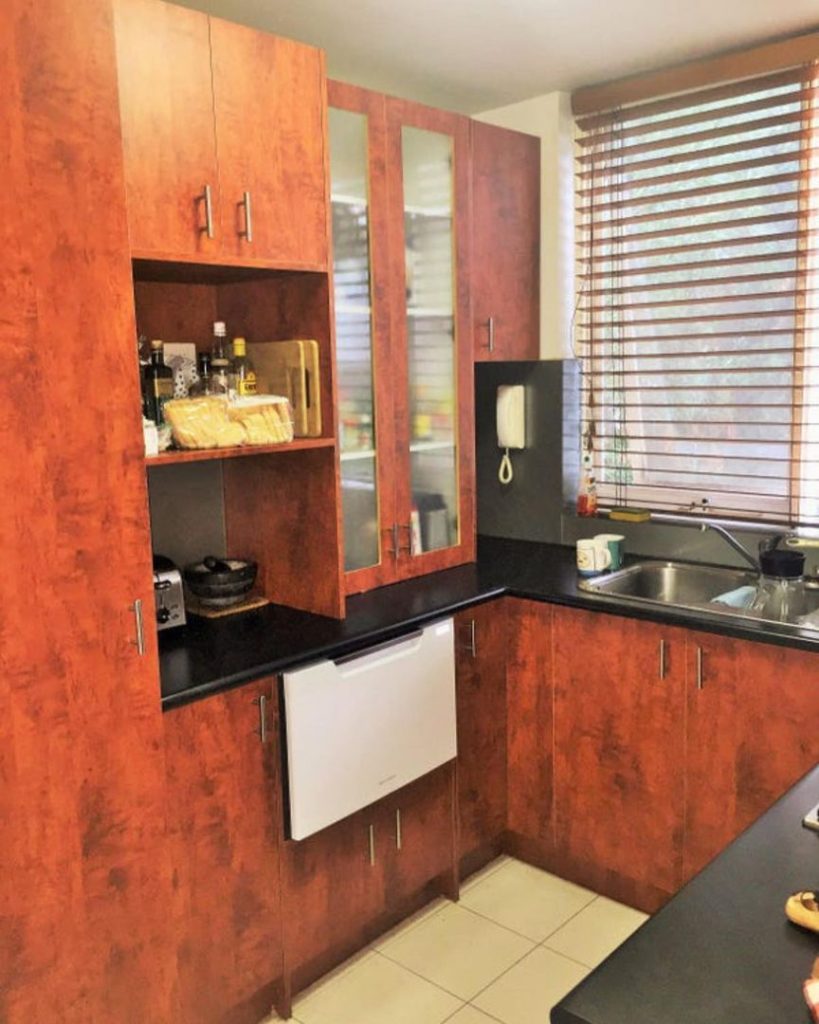 Kitchen corner with reddish-brown wooden cabinets, black countertop, integrated dishwasher, and double sink under a window with blinds.