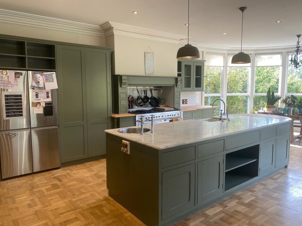 Modern kitchen with green cabinets, marble countertop island, stainless steel refrigerator, and large windows.