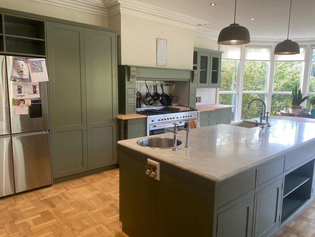 Modern kitchen with green cabinets, marble island, stainless steel refrigerator, and large windows.