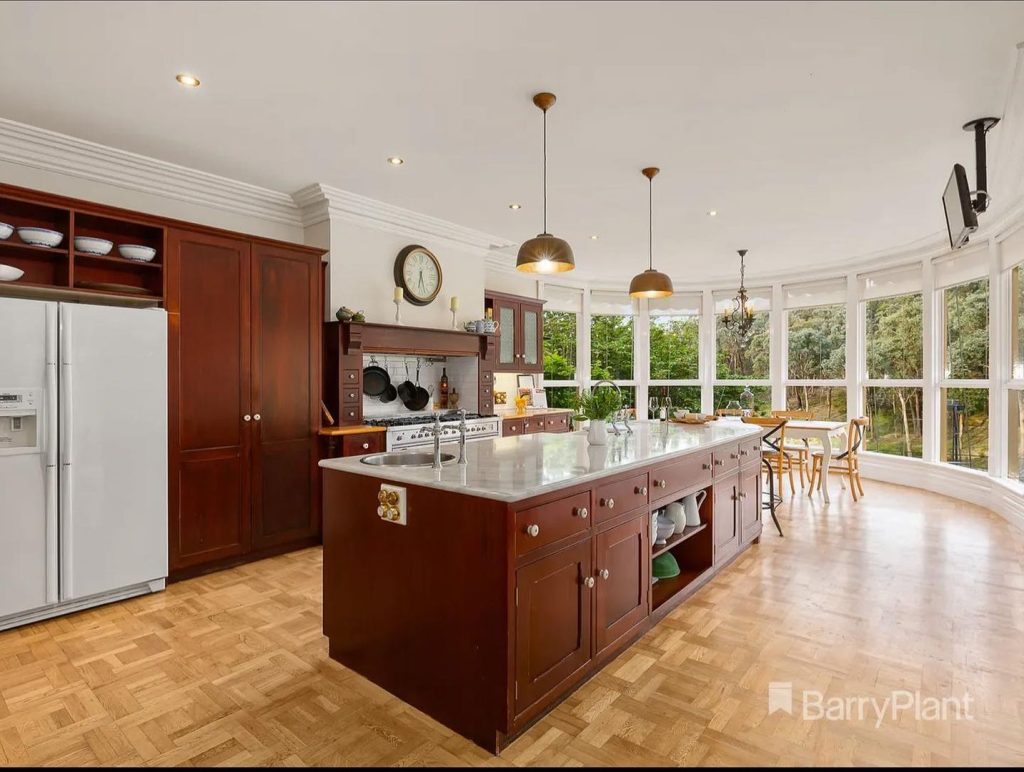 Large kitchen with wooden cabinets, marble countertops, central island, pendant lights, and dining area near large windows.