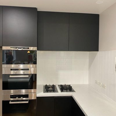 Modern kitchen with black cabinets, stainless steel ovens, gas burners, and white tiled backsplash.