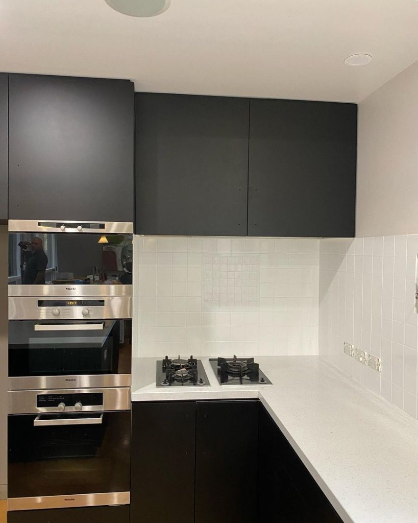 Modern kitchen with black cabinets, stainless steel ovens, gas burners, and white tiled backsplash.