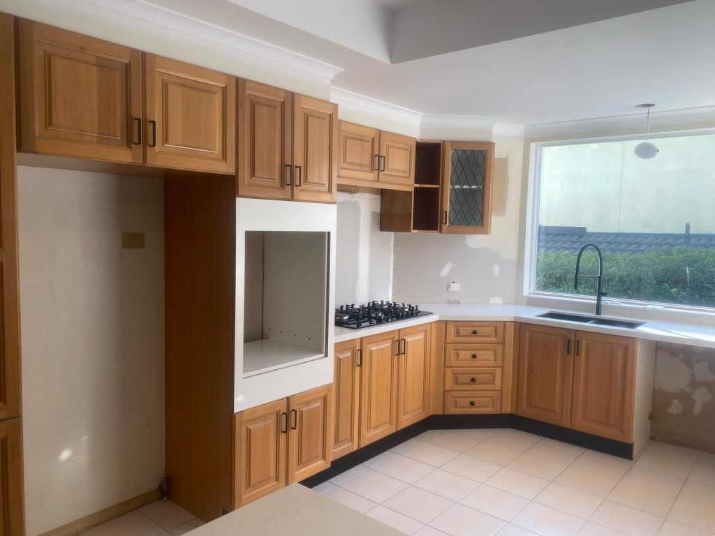 Partially renovated kitchen with wooden cabinets, gas stovetop, black faucet, and large window.