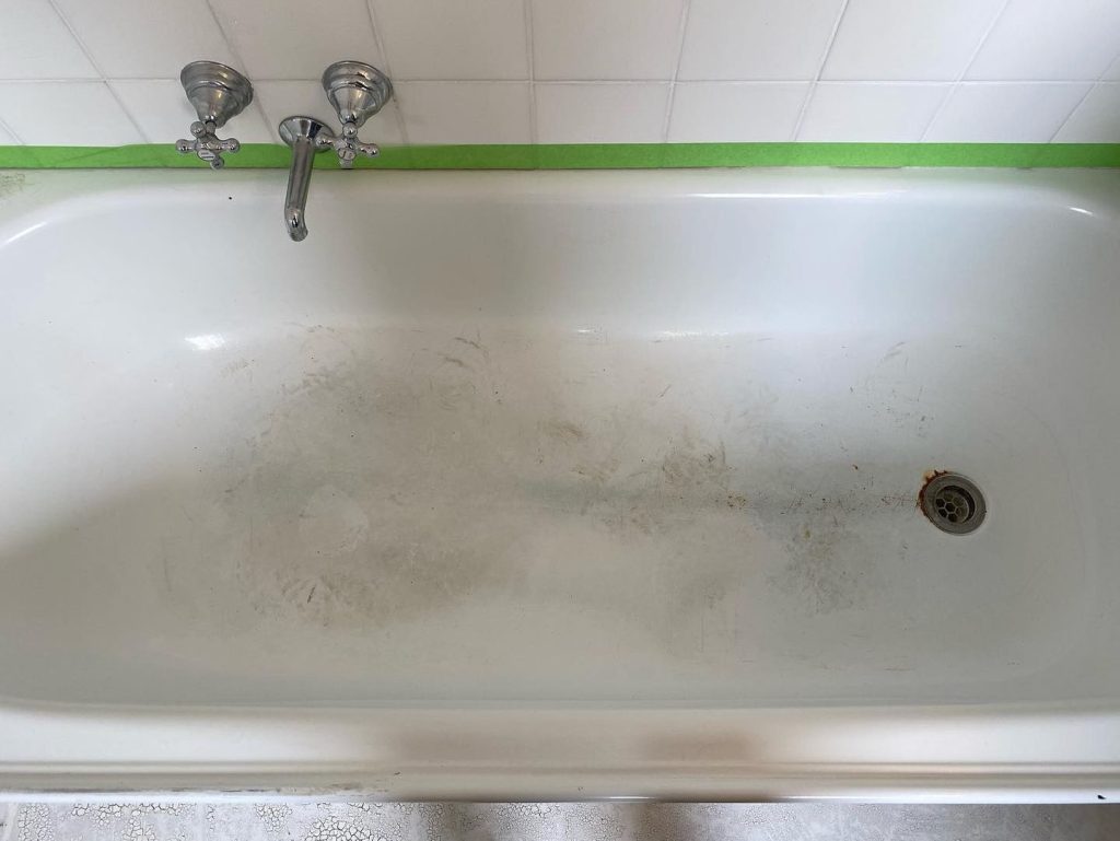 A white bathtub with dirt and stains, prepared for resurfacing with green painter's tape around the faucet area.