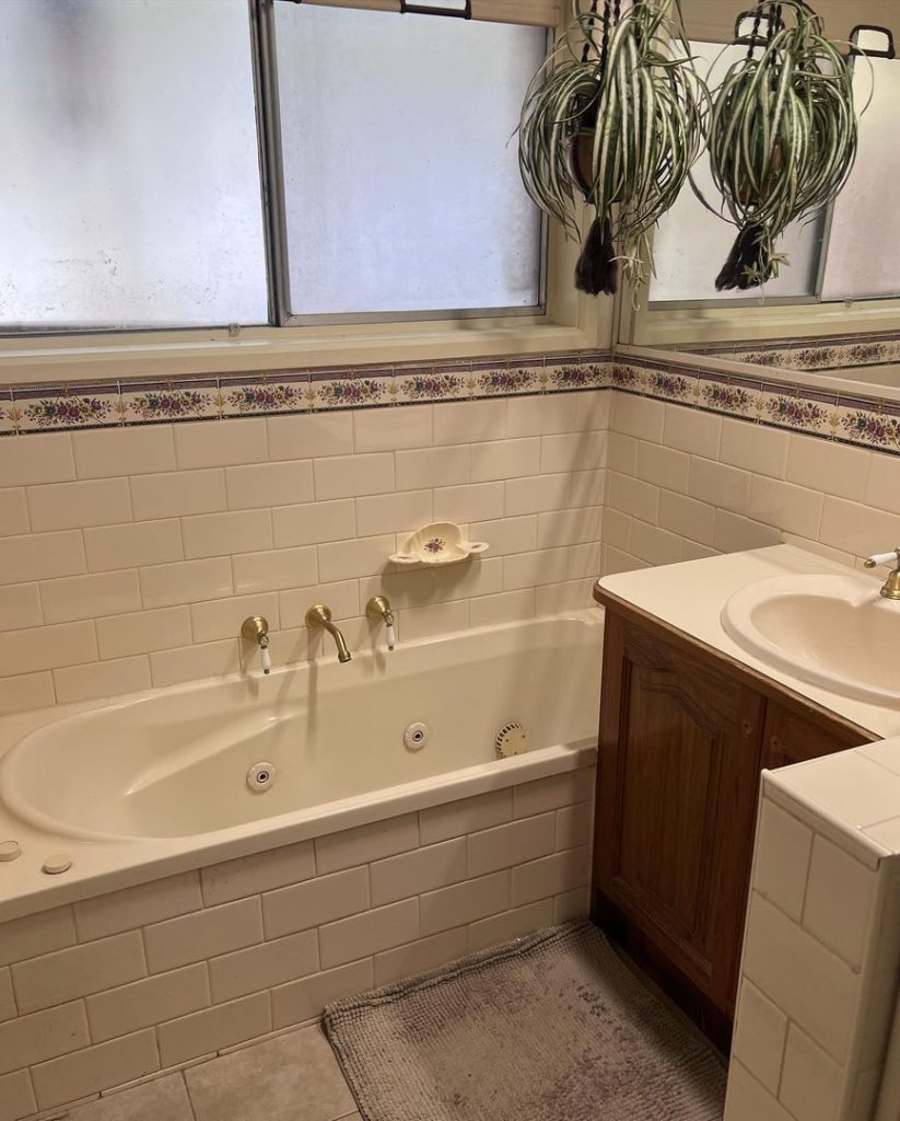 Bathroom with white tiles, floral border, bathtub with gold faucets, wooden sink cabinet, and hanging plants.