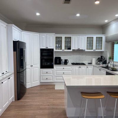 Modern kitchen with white cabinets, stainless steel refrigerator, built-in oven and microwave, gas cooktop, wooden flooring, and large window.