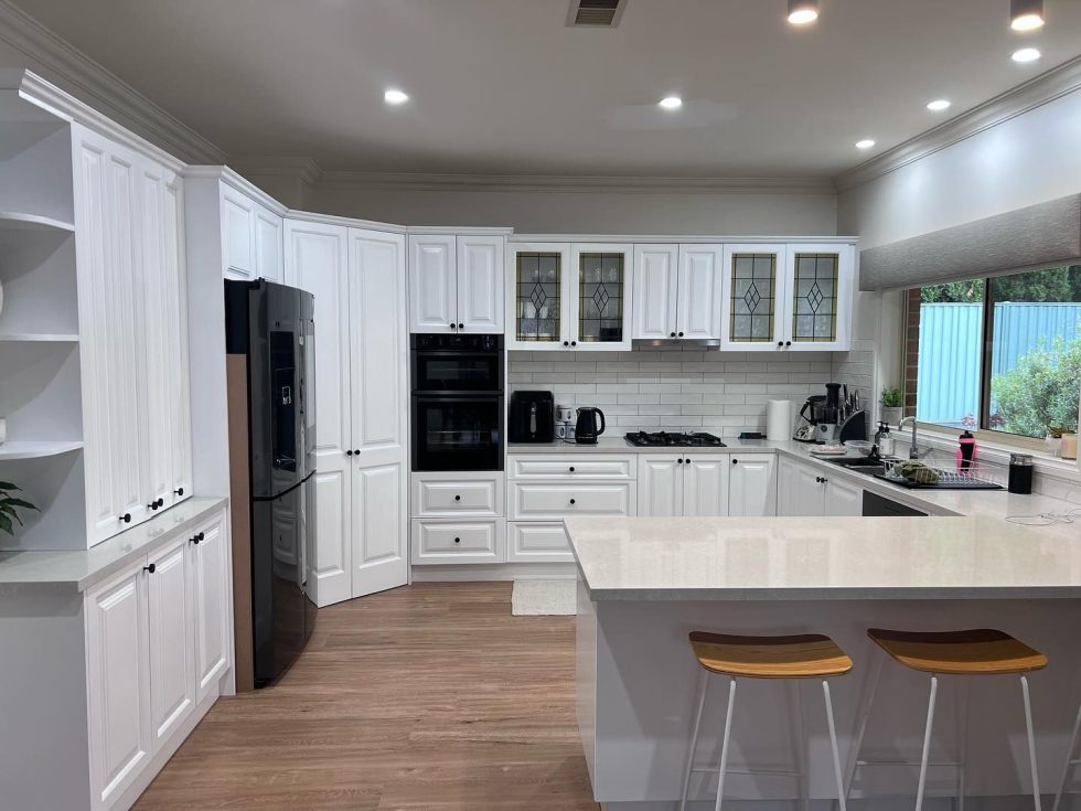 Modern kitchen with white cabinets, stainless steel refrigerator, built-in oven and microwave, gas cooktop, wooden flooring, and large window.