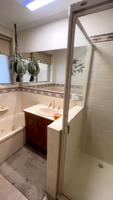 Bathroom showing a bathtub, sink with wooden cabinet, hanging plants, and glass-door shower area.