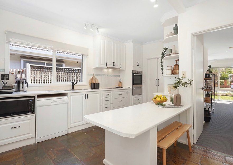 Modern white kitchen with an island countertop, slate tile flooring, and natural light from a large window.