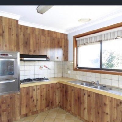 Kitchen with rustic wooden cabinets, stainless steel oven, gas stovetop, double sink under a large window, and beige countertops.