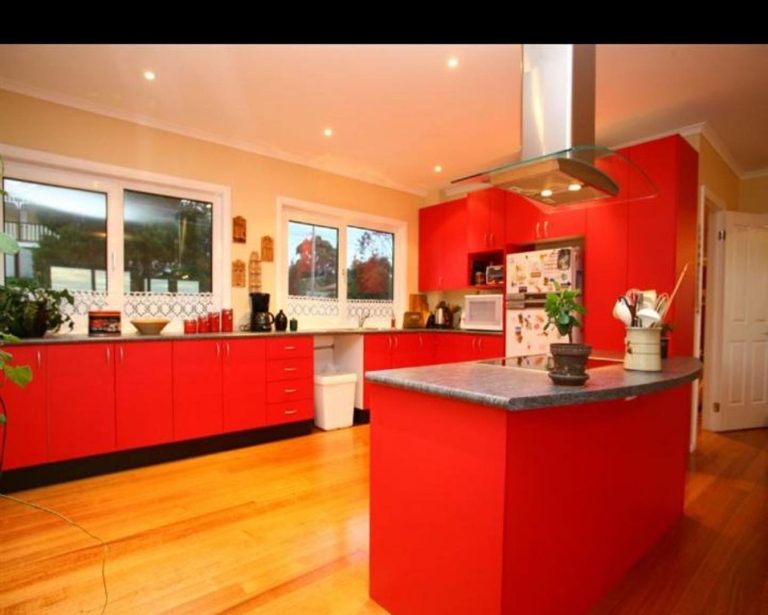 Modern kitchen with red cabinets, grey countertops, wooden flooring, and stainless steel appliances.