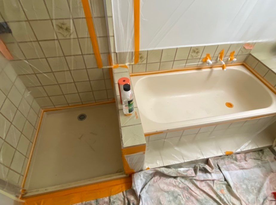 Bathroom under renovation with taped-off bathtub and shower area, plastic sheeting on tiles, and protective floor covering.