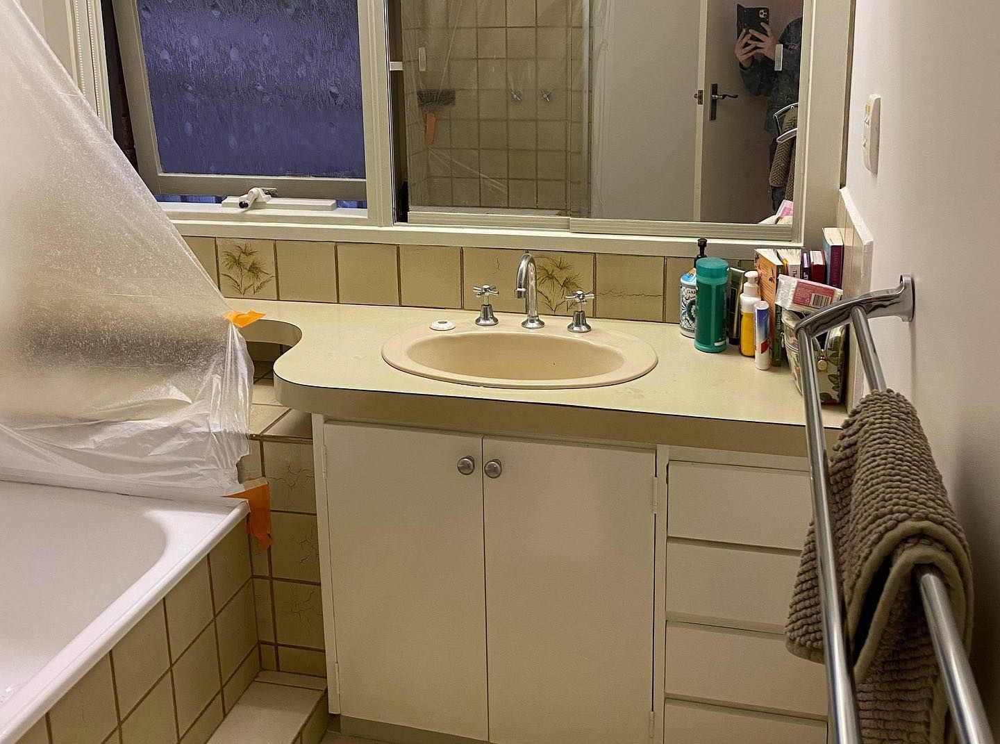 Bathroom with beige sink, plastic-covered bathtub, floral tiles, and toiletries on counter.