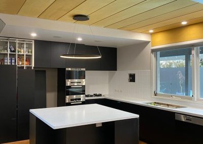 Modern kitchen with black cabinets, white countertops, central island, stainless steel appliances, and wooden ceiling panels.